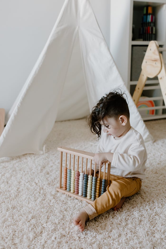 A young boy sitting on a carpeted floor playing with a colorful wooden abacus beside a white teepee tent.